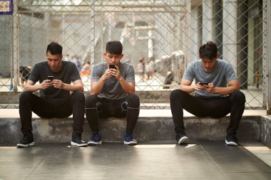 Üç genç Asyalı yetişkin adam dışarıda cep telefonlarına bakıyor.