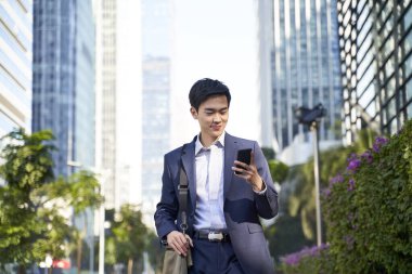Genç Asyalı işadamı modern şehrin merkezinde sokakta yürürken cep telefonundan mesajlara bakıyor.