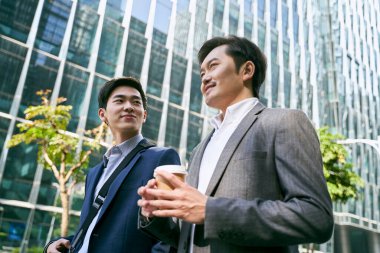 İki Asyalı beyaz yakalı ofis çalışanının modern şehrin merkezinde sokakta yürürken iş konuşmalarının yan görüntüsü.