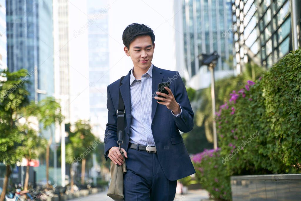 young asian businessman looking at messages on cellphone while walking in the street in downtown of modern city