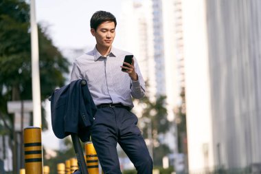 Genç Asyalı işadamı elektrikli scooterının yanında duruyor ve modern şehrin aşağısındaki caddede cep telefonuna bakıyor.