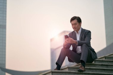 Asyalı şirket yöneticisi merdivenlerde oturmuş cep telefonuna bakıyor.