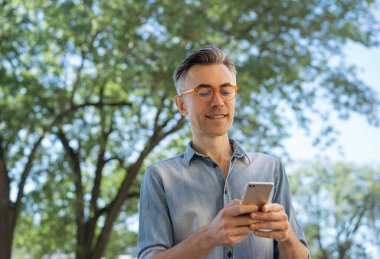 Akıllı telefon, internet, iletişim, sohbet, online alışveriş yapan, parkta duran orta yaşlı bir adam. Yakışıklı, olgun iş adamının portresi cep telefonu tutuyor, internetten haberleri okuyor. 