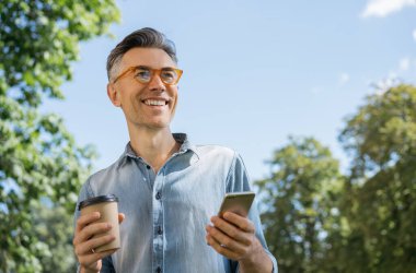 Elinde bir fincan kahve ve akıllı telefon ile parkta yürüyen şık gözlüklü, gülümseyen olgun bir adamın portresi. Çevrimiçi alışveriş için mobil uygulama kullanan orta yaşlı hipster 