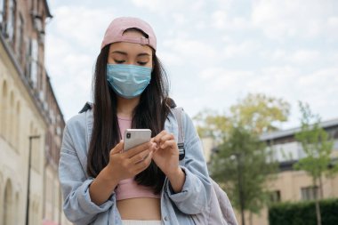 Korunan tıbbi maske takan Asyalı bir kadın cep telefonu kullanıyor, internetten iletişim kuruyor, mesajlaşıyor, sokakta yürüyor. Karantina, virüs, güvenli konsept