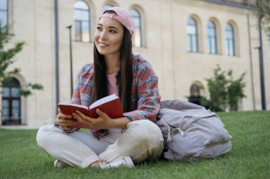 Üniversite kampüsünde okuyan, kitap okuyan, sınav hazırlığı yapan genç ve çekici bir öğrenci. Düşünceli Asyalı kadın portresi. Çimlerin üzerinde günlük giysiler giyiyor. Eğitim kavramı