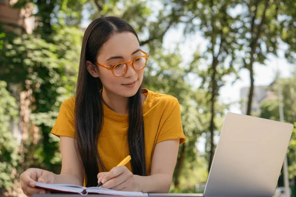 Asian student studying, exam preparation, distance learning, online education concept. Smiling woman using laptop computer, watching training courses, taking notes, working outdoors