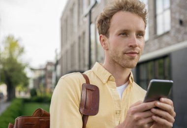 Genç yakışıklı iş adamı günlük kıyafetler giyiyor ve kurye çantası takıyor cep telefonuyla sokakta yürüyor. Çevrimiçi alışveriş yapan başarılı adamın portresi, mobil bankacılık konsepti