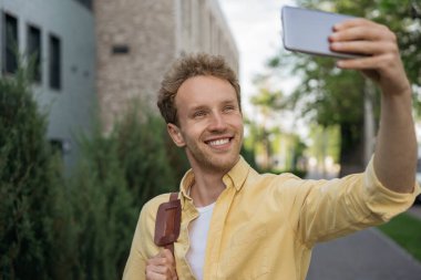 Sarı tişört giyen gülümseyen genç adamın portresi cep telefonunda selfie çekiyor. Kendine güvenen, video yayınlayan, sokakta iletişim kuran biri.