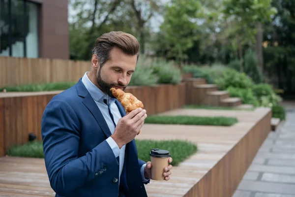 Young handsome businessman holding cup of coffee, eating croissant on the street. Happy bearded man holding tasty food sitting in park, lunch time, coffee break concept