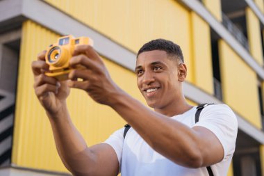 Afro-Amerikalı mutlu fotoğrafçının sokakta fotoğraf çekerken, başka tarafa bakarken portresi. Sarı dijital kameralı gülümseyen genç turist, açık havada fotoğraf çekiyor. İlham, seyahat konsepti