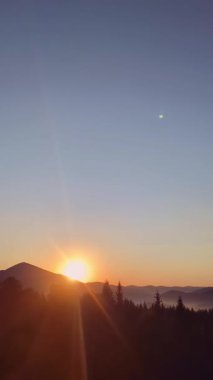 Dağların tepelerinde ve siluetli orman ağaçlarının üzerinde çarpıcı bir güneş parlayan renkli bir güneşin nefes kesici dikey görüntüsü.