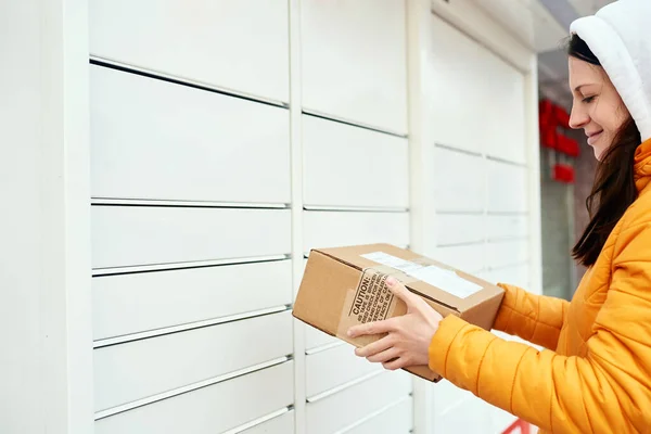 La ragazza manda il pacco alla posta nell'armadietto. Ricevimento pacco. La donna tiene una scatola, guarda il pacco e sorride. — Foto Stock