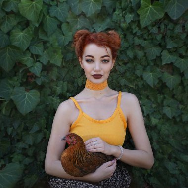 A beautiful red-haired young woman holds a brown hen in her arms against a background of emerald green ivy. Artistic processing. clipart