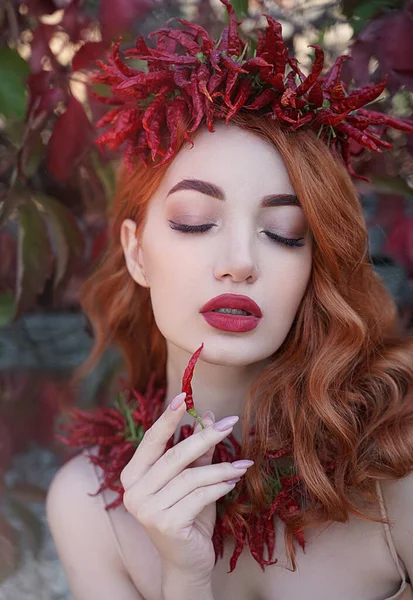 Retrato de una mujer sexy pelirroja con los labios llenos de rojo en una corona y un collar de chiles picantes con un pimiento en las manos. —  Fotos de Stock