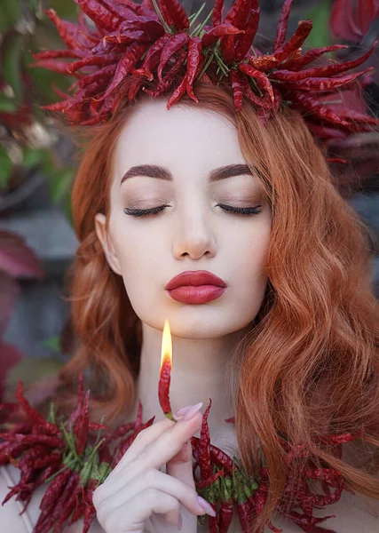 Retrato de uma mulher sexy de cabelos vermelhos com lábios vermelhos gordos em uma coroa e um colar de pimentas quentes segurando uma pimenta. — Fotografia de Stock