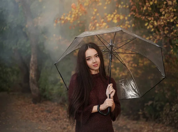 Joven mujer pensativa bajo un paraguas transparente sobre un fondo de otoño y humo. —  Fotos de Stock