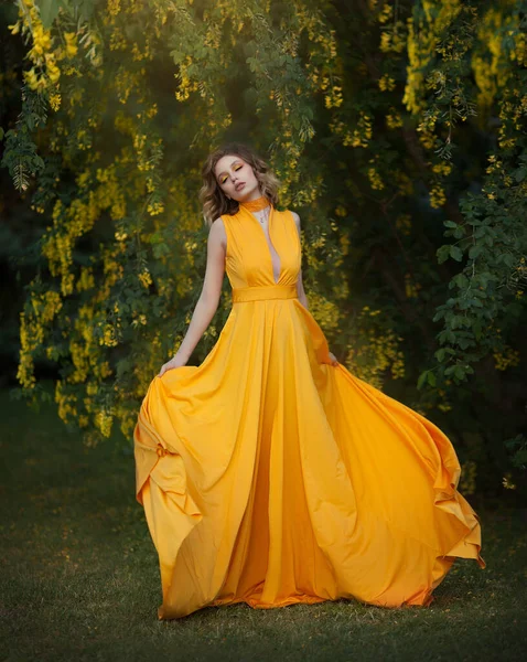 Donna in abito lungo giallo svolazzante sotto un albero in fiore con fiori gialli, stoffa di seta ondulante, lavorazione artistica. — Foto Stock