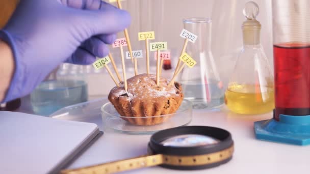 Conceito de alimentação saudável. A mão do assistente de laboratório está segurando uma lupa em suas mãos, o cupcake na mesa é decorado com comprimidos com os nomes dos aditivos de E. Alimentos — Vídeo de Stock