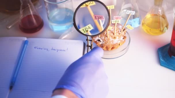 Healthy eating concept. The hand of the laboratory assistant is holding a magnifier in his hands, the cupcake on the table is decorated with tablets with the names of the additives of E. Food — Stock Video