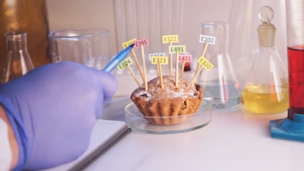 Concept d'alimentation saine. La main de l'assistant de laboratoire tient une loupe, un cupcake décoré de comprimés avec les noms des additifs E. Laboratoire alimentaire. — Video
