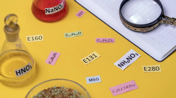 Signature with the name of the food additive under a magnifying glass on a table with flasks and a notebook. Unhealthy food concept. — Foto de Stock