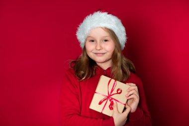 Noel Baba şapkası ve kırmızı kazağı olan güzel genç kız kırmızı arka plan önünde bir hediye paketi tutuyor.