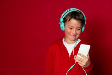 Turkuaz kulaklıklı, beyaz akıllı telefonlu ve kırmızı kazaklı havalı bir çocuk.