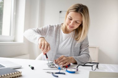Ofisteki masada oturan sarışın kadın elektrikçi prizi tamir ediyor.