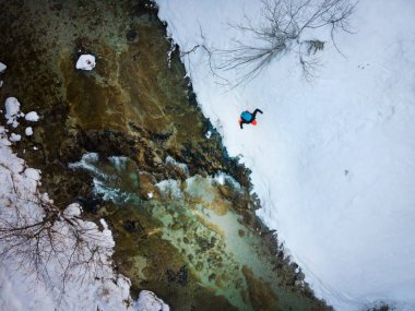 Sırt çantalı, turuncu şapkalı, turuncu pantolonlu, kar ve buzun içinde soğuk dağ nehrinin yanında tehlikeli bir yolda yürürken drone görüntüsü.