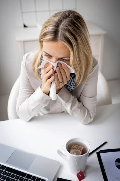 Sick businesswoman in office feeling unwell sitting in front of her laptop blowing her nose, stressed femal employee have anxiety attack at workplace