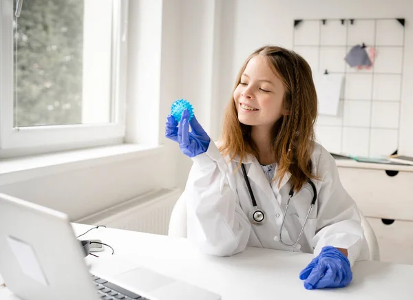 pretty blonde and young schoolgirl girl plays doctor and has white coat on and blue protective gloves and watches a blue ball, a model of a virus
