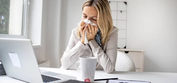 Sick business woman in office feeling unwell sitting in front of her laptop blowing her nose, stressed female employee have anxiety attack at workplace