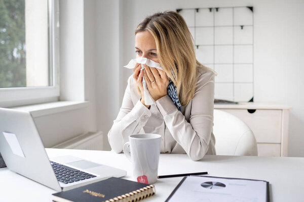 Sick business woman in office feeling unwell sitting in front of her laptop blowing her nose, stressed female employee have anxiety attack at workplace