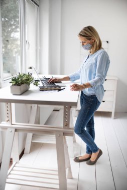 ffp2 maskeli bir iş kadını dizüstü bilgisayarla çalışıyor, masasında duruyor..