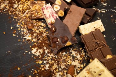 Bir sürü çikolata çeşidi ve fındık ve kakao tozu gibi çeşitli malzemeler yukarıdan çekilen siyah bir tahta tabakta duruyor.