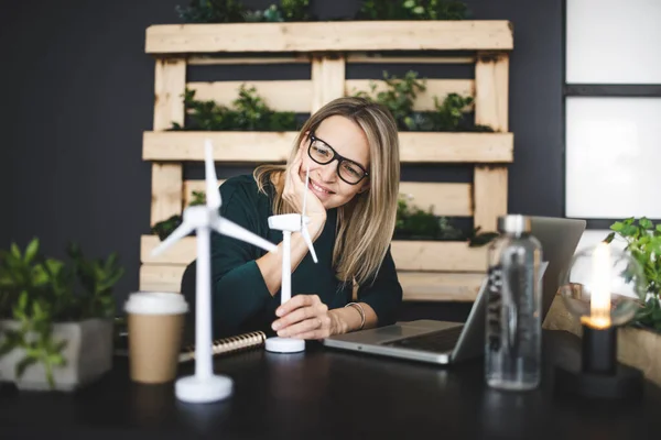 pretty, young woman with stylish, modern black glasses sits in a sustainable office and works with wind turbine model