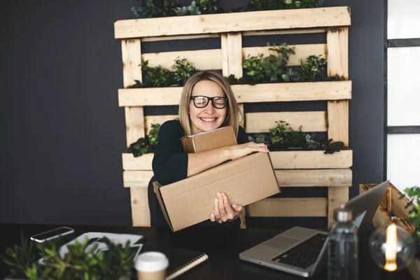 blonde woman with brown parcels is sitting in front of laptop in a very modern, ecological, sustainable office and is happy about her online order