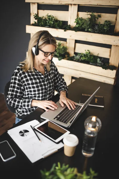 pretty, young blond businesswoman with black and white checked shirt is sitting in a sustainable, ecological office with a headset and is having a web meeting