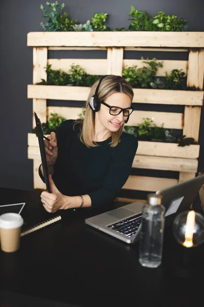 pretty, young, blond woman with stylish, modern black glasses sits in a sustainable office and has an online web meeting with a headset