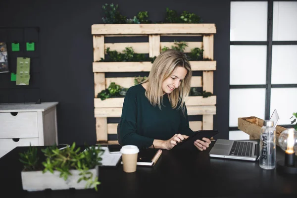 pretty young blond woman is sitting in a modern, sustainable office with lots of green ecological plants and is working on her table