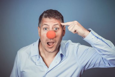 Orta yaşlı, mavi gömlekli ve kırmızı burunlu bir adam stüdyoda mavi arka planda poz veriyor.