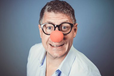 Mavi gömlekli, siyah gözlüklü ve kırmızı burunlu bir işadamı stüdyoda mavi arka planda poz veriyor.