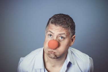 Orta yaşlı, mavi gömlekli ve kırmızı burunlu bir adam stüdyoda mavi arka planda poz veriyor.