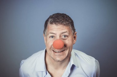 Orta yaşlı, mavi gömlekli ve kırmızı burunlu bir adam stüdyoda mavi arka planda poz veriyor.