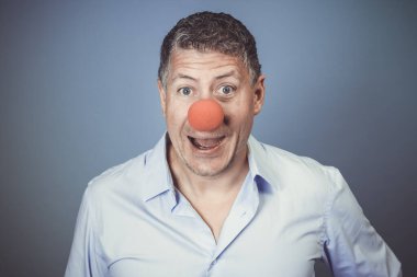 Orta yaşlı, mavi gömlekli ve kırmızı burunlu bir adam stüdyoda mavi arka planda poz veriyor.