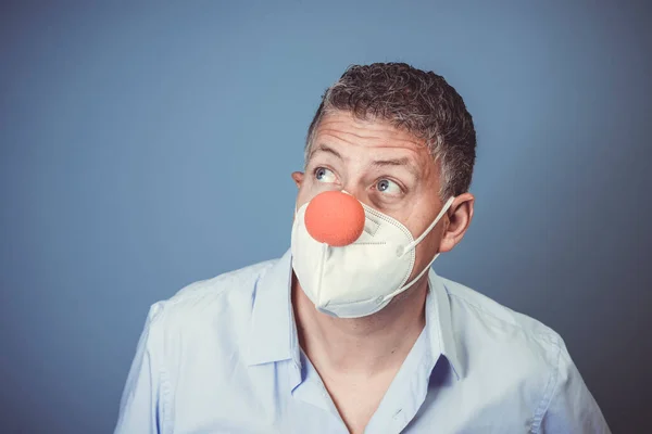 Middle aged man with nose mouth protection and red clown nose in front of blue background