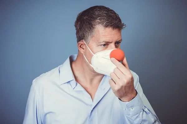 Middle aged man with nose mouth protection and red clown nose in front of blue background