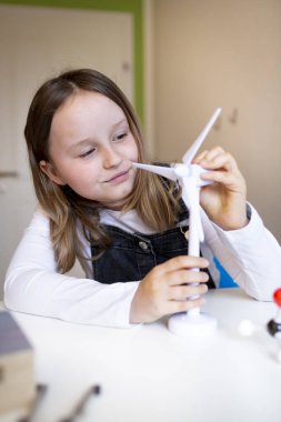 Güzel liseli kız evdeki masasında oturmuş evde eğitim görürken rüzgar türbininin modeliyle oynuyor.