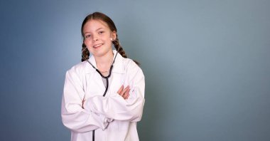 İki örgülü güzel kız doktor gibi giyinmiş ve steteskopla mavi arka planda poz veriyor.